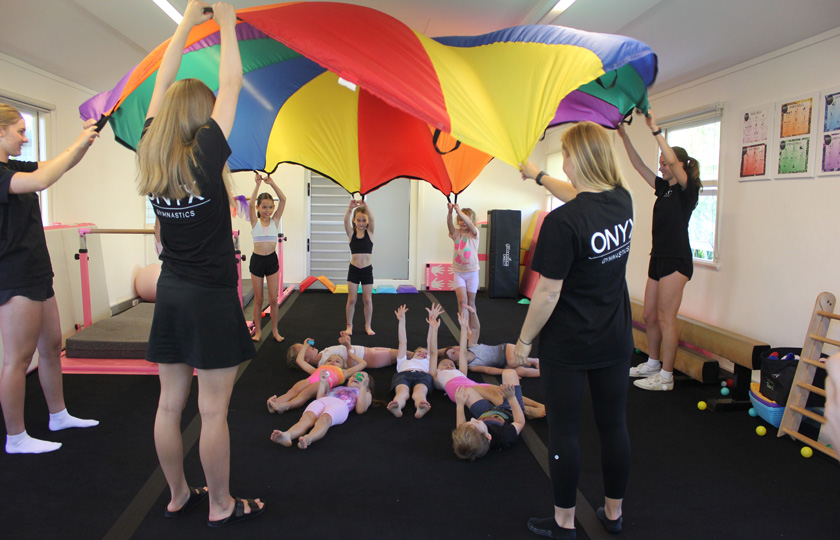 Children lying on the floor during an Onyx Gymnastics class while instructors hold a rainbow parachute above them.